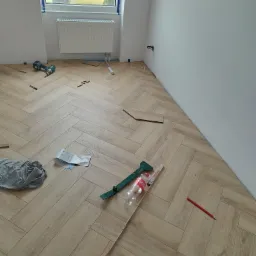 Układanie parkietu w jodełkę francuską, widoczne narzędzia, niedopita butelka Coca-Coli, ołówek stolarski i fragment instrukcji, w tle okno z widokiem na budynek.