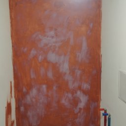 MaxBud - Ściana w trakcie malowania na kolor terakoty, z widocznymi smugami i niedomalowaniami, instalacja wodna z rurami w kolorach czerwonym i niebieskim po prawej stronie.