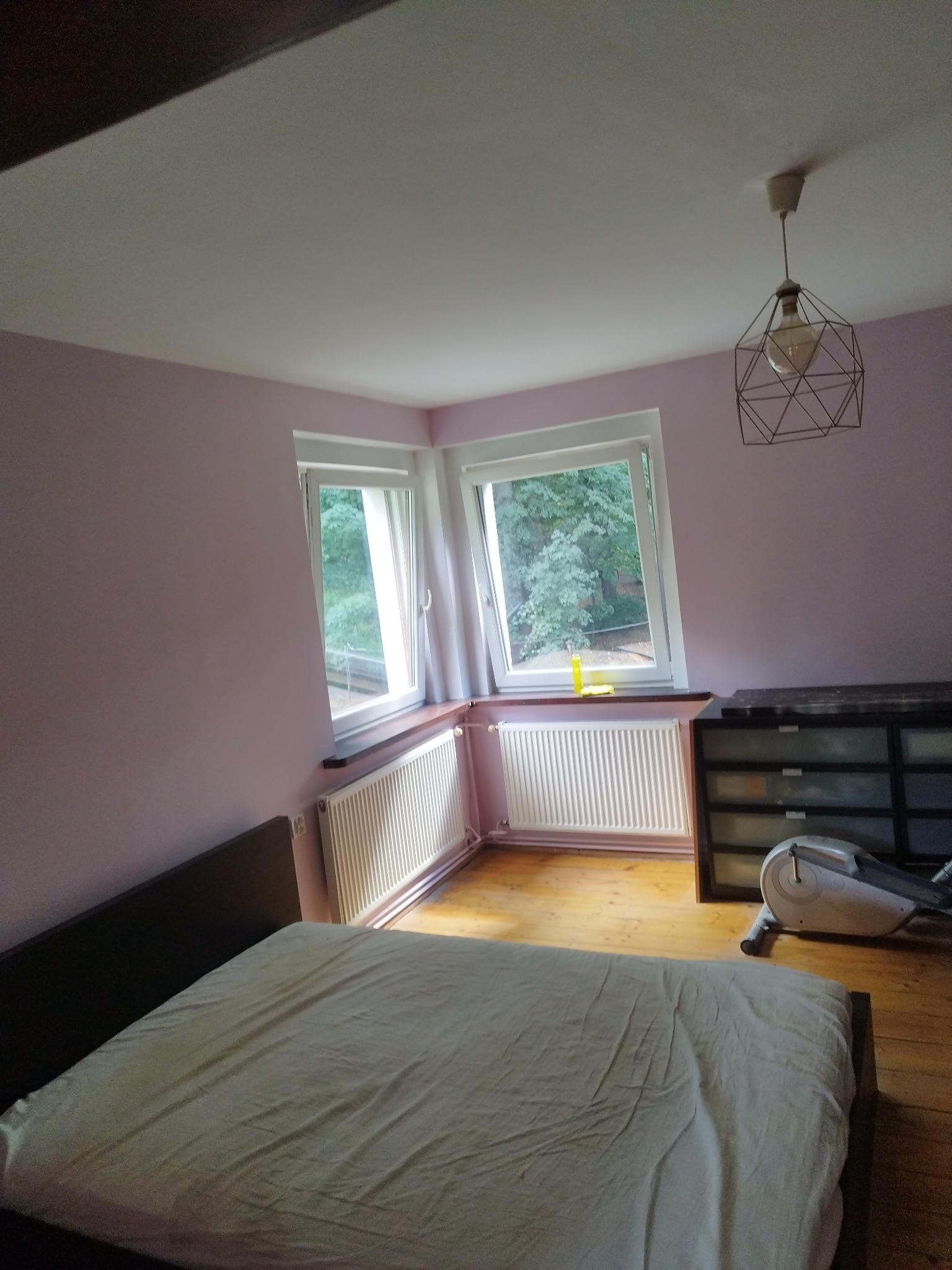 Pomieszczenie z łóżkiem, ścianami pomalowanymi na liliowo, dwoma oknami z widokiem na zieleń, grzejnikami, drewnianą podłogą i geometryczną lampą wiszącą. W tle widoczny sprzęt do ćwiczeń.