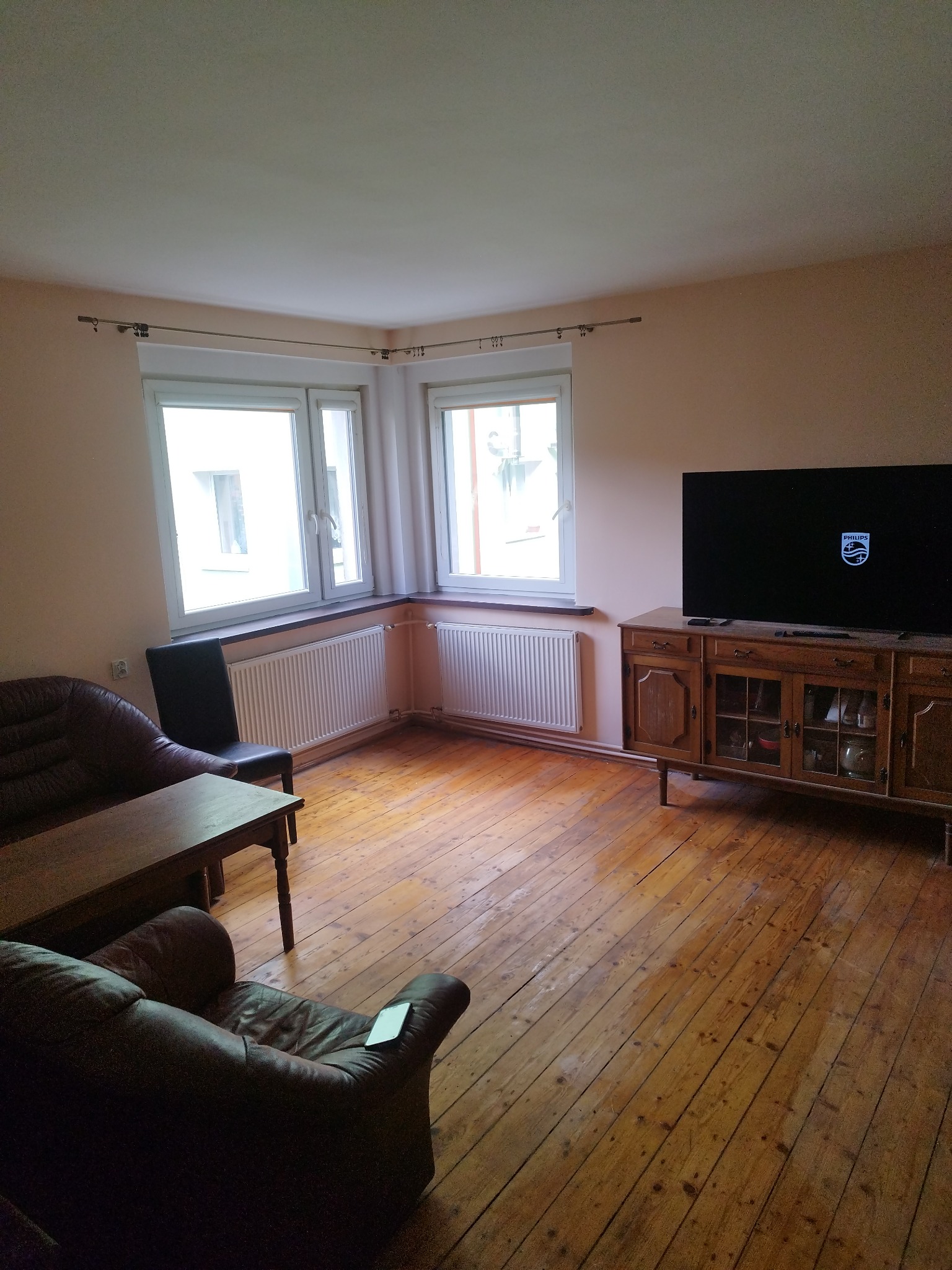 Pokój dzienny z drewnianą podłogą, brązowymi meblami, telewizorem i dwoma oknami narożnymi z białymi ramami.