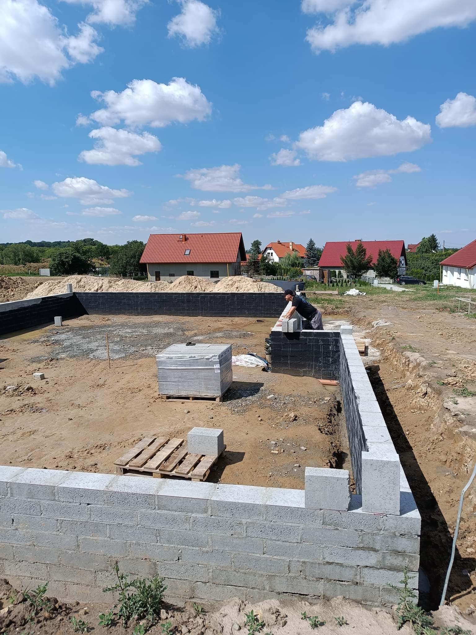 Wznoszenie fundamentów z bloczków betonowych na tle zabudowy jednorodzinnej i błękitnego nieba z chmurami.