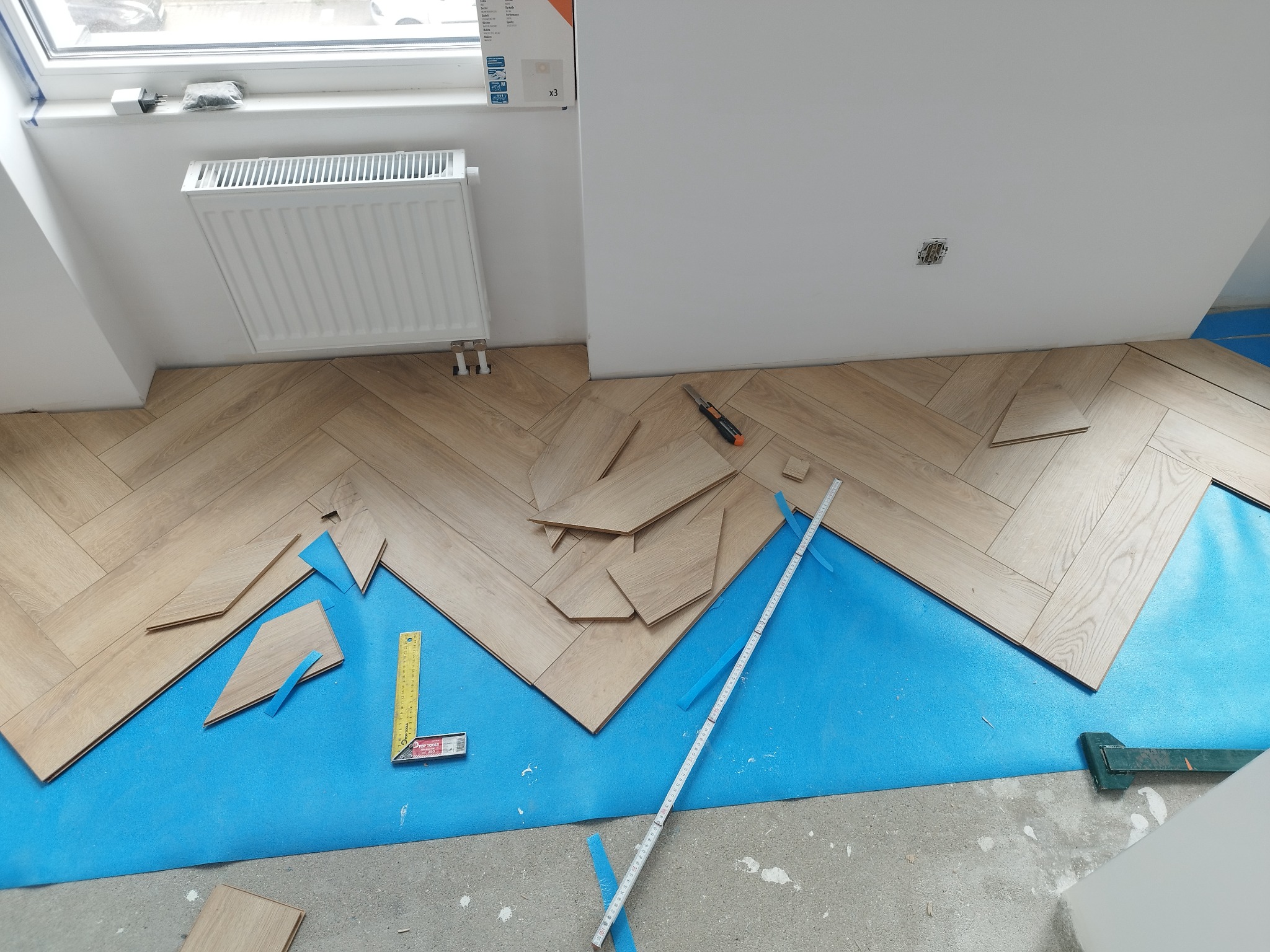 Układanie jasnych paneli podłogowych w jodełkę na niebieskim podkładzie, widoczne narzędzia: miarka, nóż i kątownik.