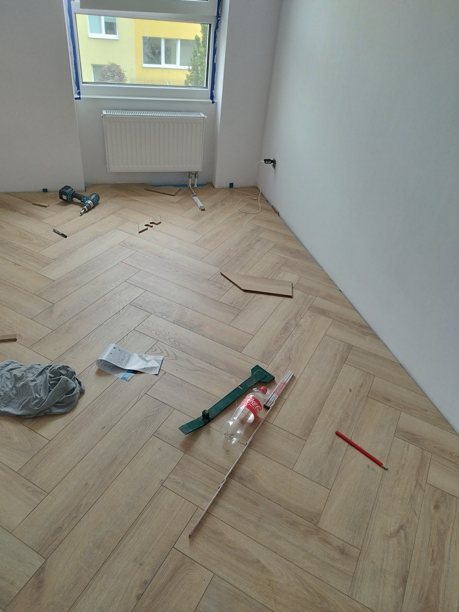 Układanie parkietu w jodełkę francuską, widoczne narzędzia, niedopita butelka Coca-Coli, ołówek stolarski i fragment instrukcji, w tle okno z widokiem na budynek.