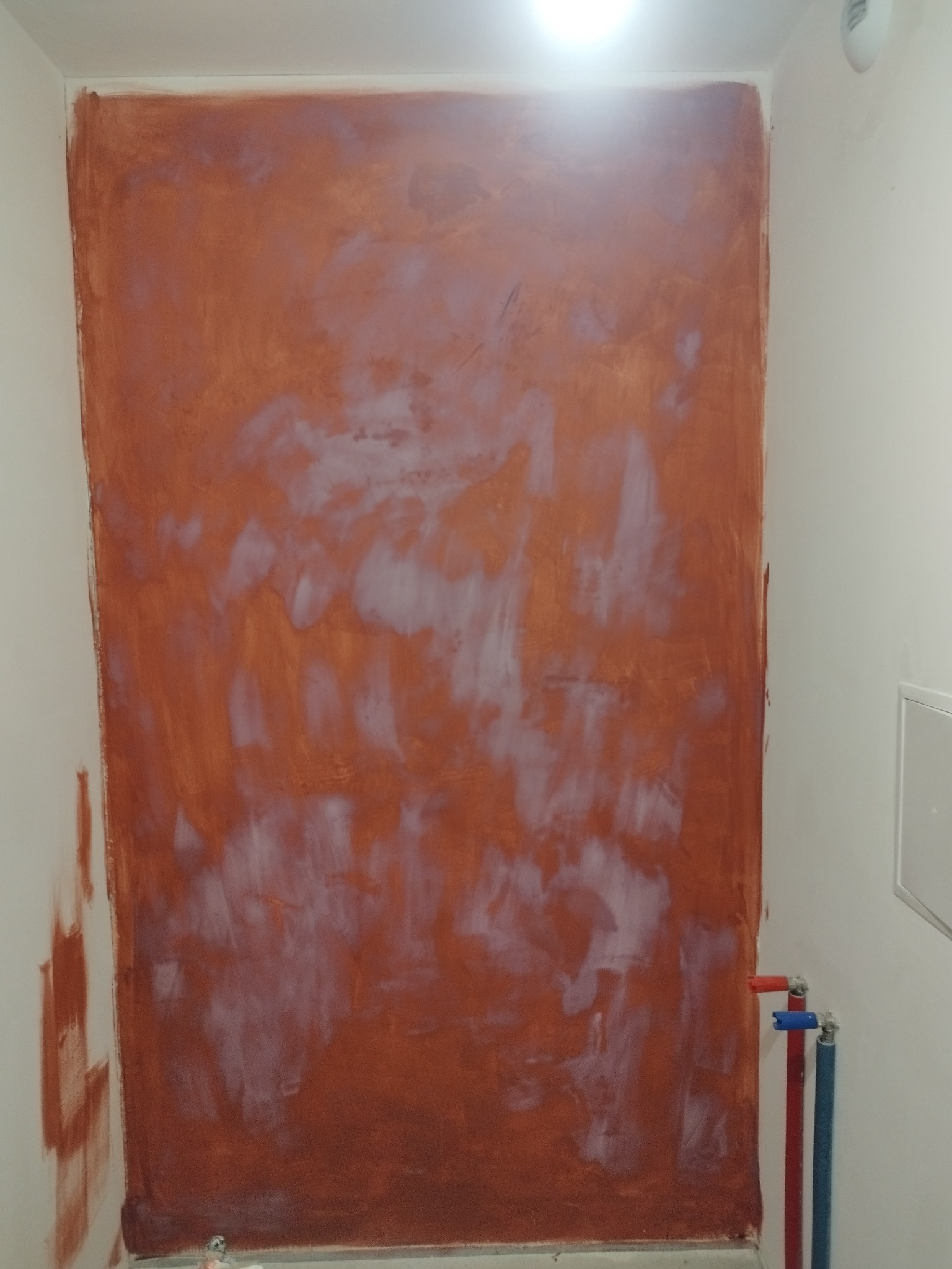 Ściana w trakcie malowania na kolor terakoty, z widocznymi smugami i niedomalowaniami, instalacja wodna z rurami w kolorach czerwonym i niebieskim po prawej stronie.