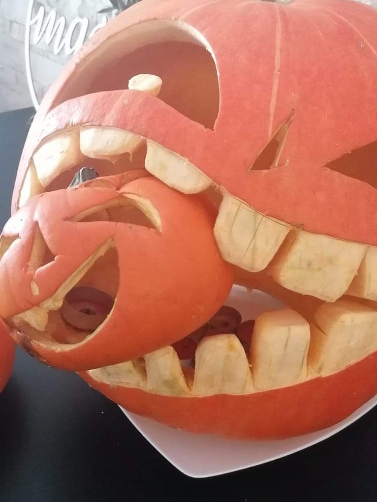 Rzeźba z dyni na Halloween przedstawiająca dużą dynię z wyciętymi zębami, połykającą mniejszą dynię z wyciętą buzią, na białym talerzu.
