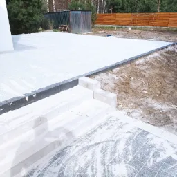 Wykończona powierzchnia z jasnoszarej kostki brukowej z jasnymi fugami, prowadząca do schodów z białego kamienia; widoczny fragment budynku z oknem, w tle ogrodzenie z drewnianych desek i blaszany...