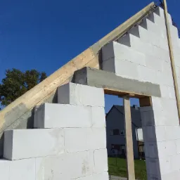 Wznoszenie ścian domu z betonu komórkowego Ytong, widoczne zbrojenie wieńca i otwór okienny z drewnianym szalunkiem, w tle błękitne niebo i fragment sąsiedniego budynku.