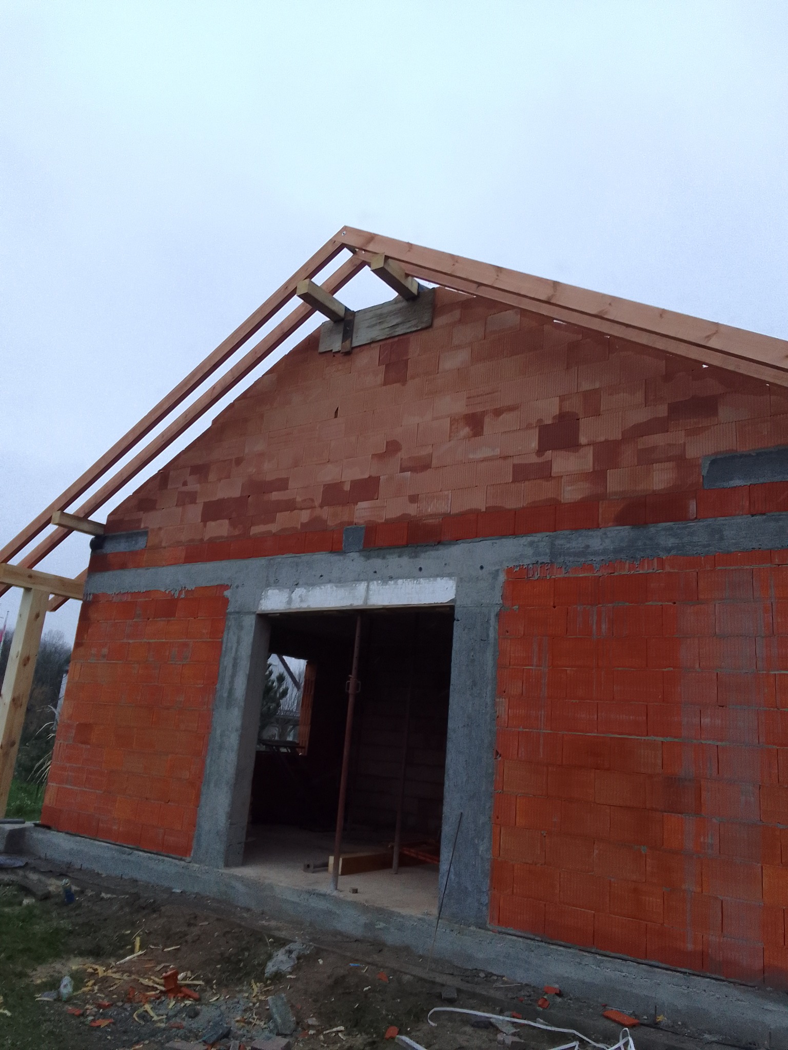 Wznoszenie ścian z czerwonej cegły ceramicznej z widocznym zbrojonym nadprożem i elementami konstrukcji dachu z drewna na tle pochmurnego nieba.