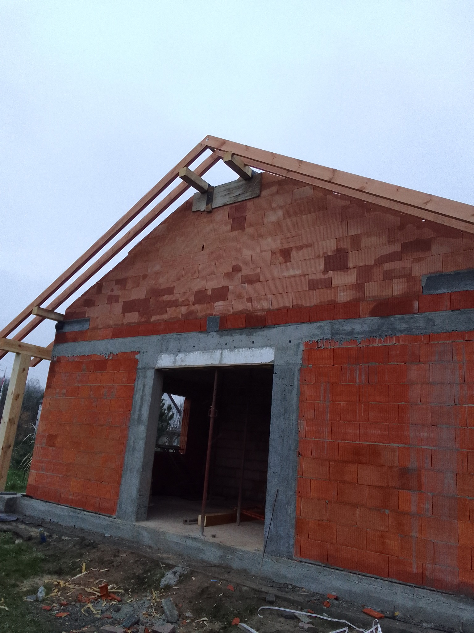 Surowy stan budynku z czerwonej cegły z widocznym szkieletem dachu z drewna, betonową ramą okienną i fundamentami, sfotografowany w pochmurny dzień.