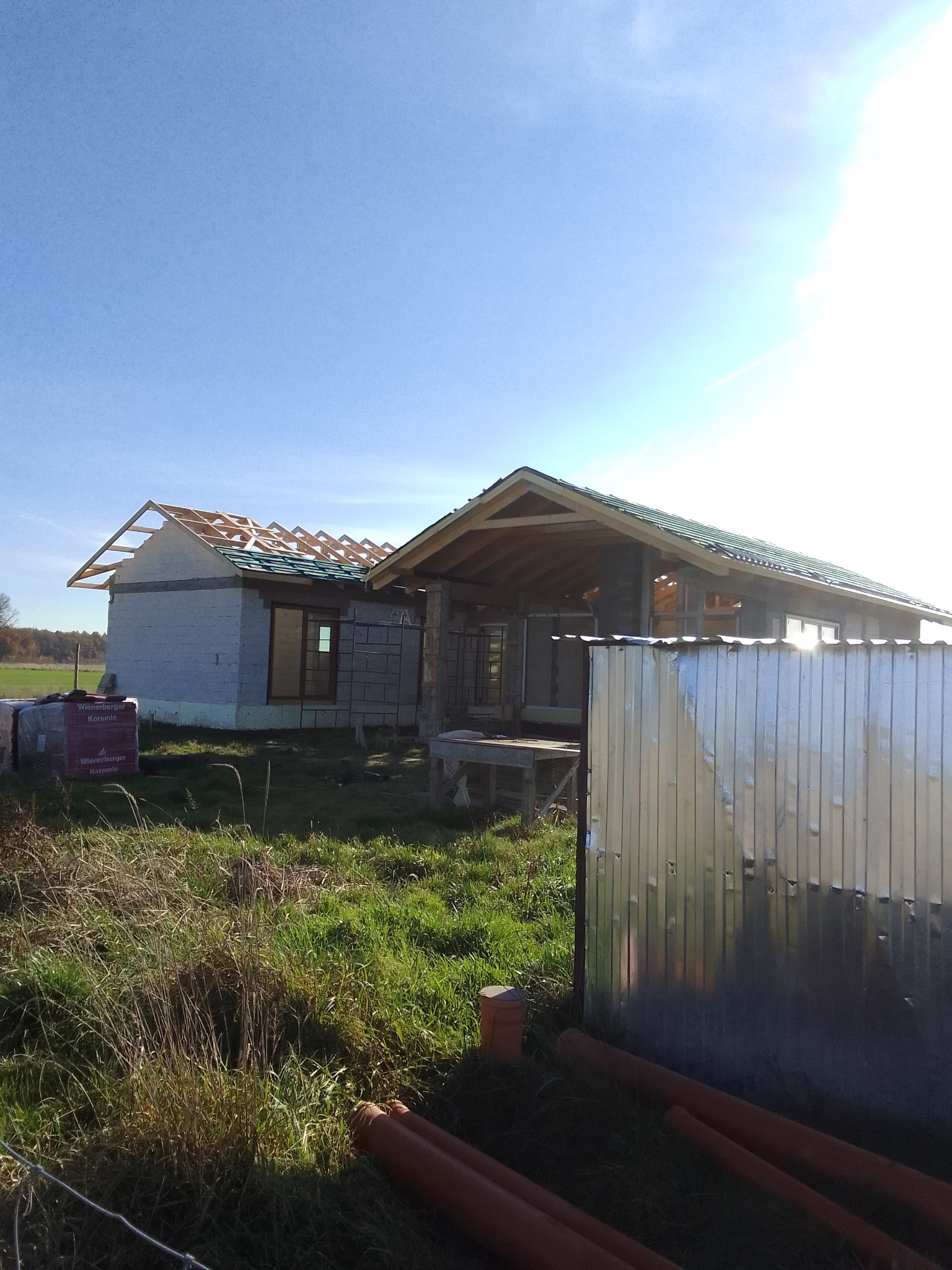 Wznoszenie dwóch budynków w stanie surowym otwartym, jeden z częściowo położonym dachem, drugi z konstrukcją dachu w trakcie budowy, widoczne materiały budowlane i tymczasowe ogrodzenie z blachy...