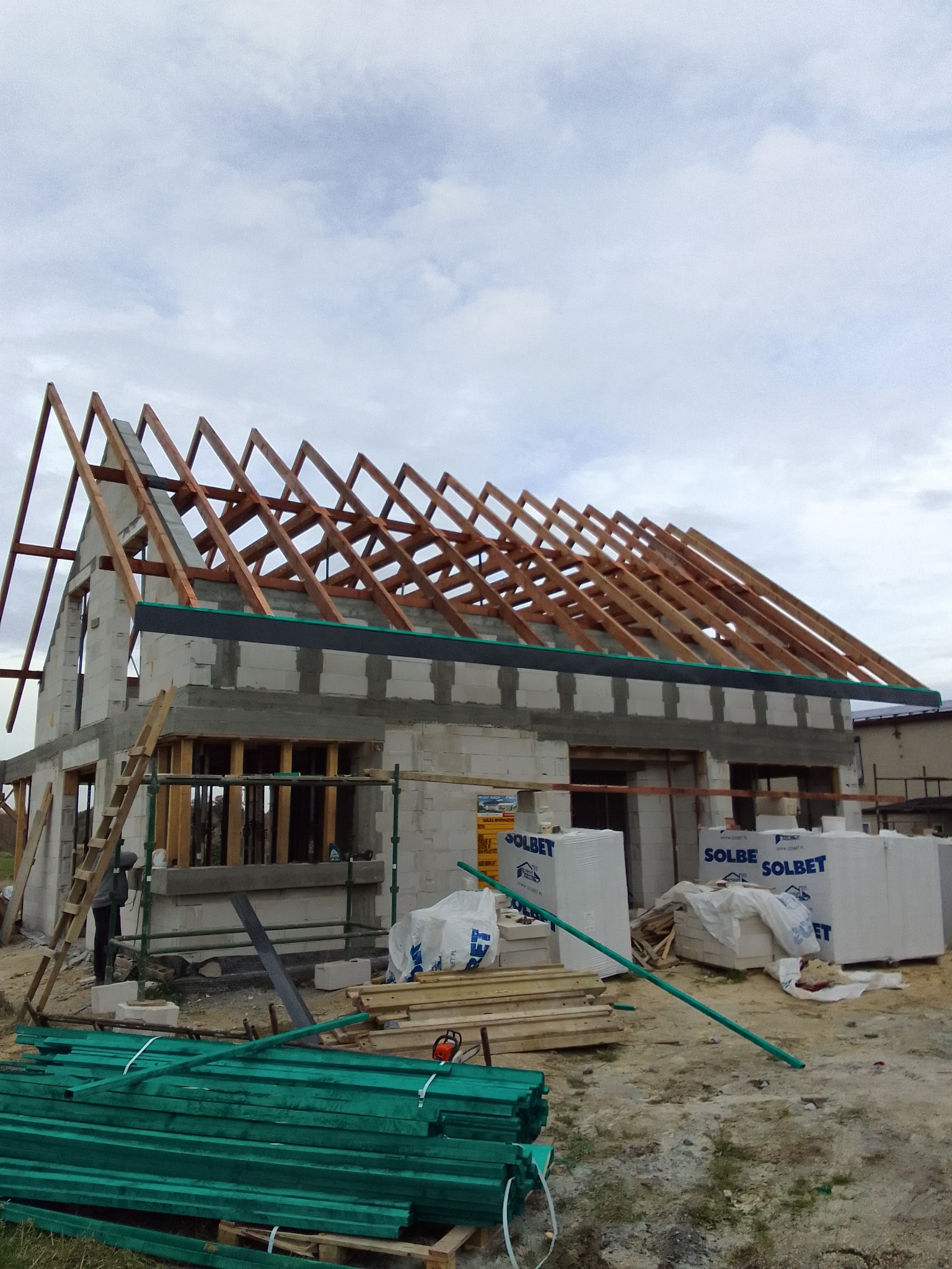 Budowa domu murowanego w trakcie realizacji: wzniesione ściany z pustaków, drewniana konstrukcja dachu, materiały budowlane składowane obok, drabina oparta o ścianę.