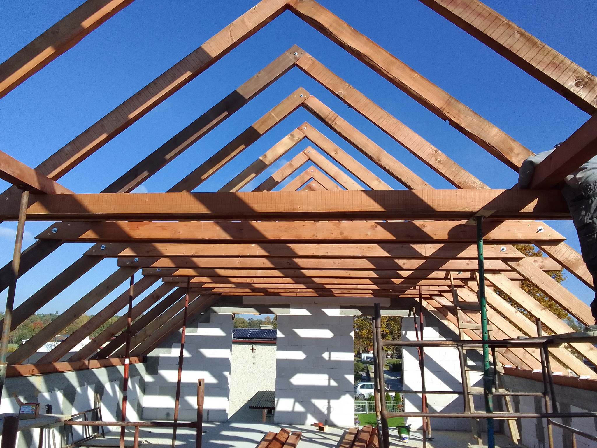 Konstrukcja drewniana dachu z widocznymi wiązarami i łatami, na tle błękitnego nieba i częściowo wymurowanych ścian budynku.