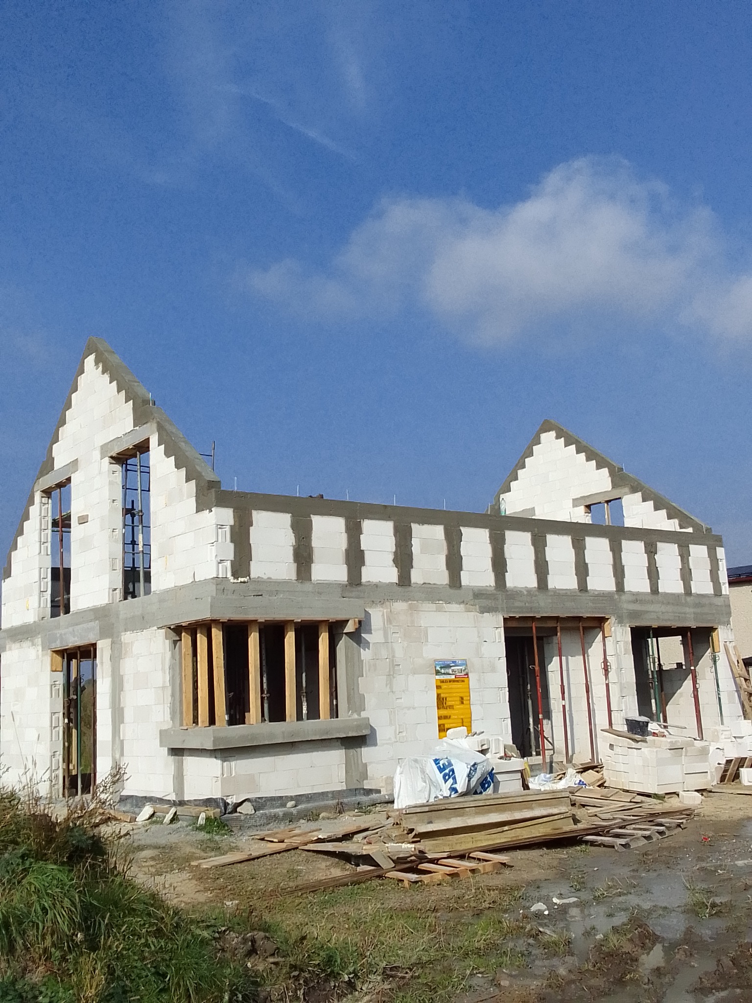 Surowy stan dwukondygnacyjnego budynku z białych bloczków, z widocznymi otworami okiennymi i elementami konstrukcyjnymi dachu, na tle błękitnego nieba z delikatnymi chmurami.