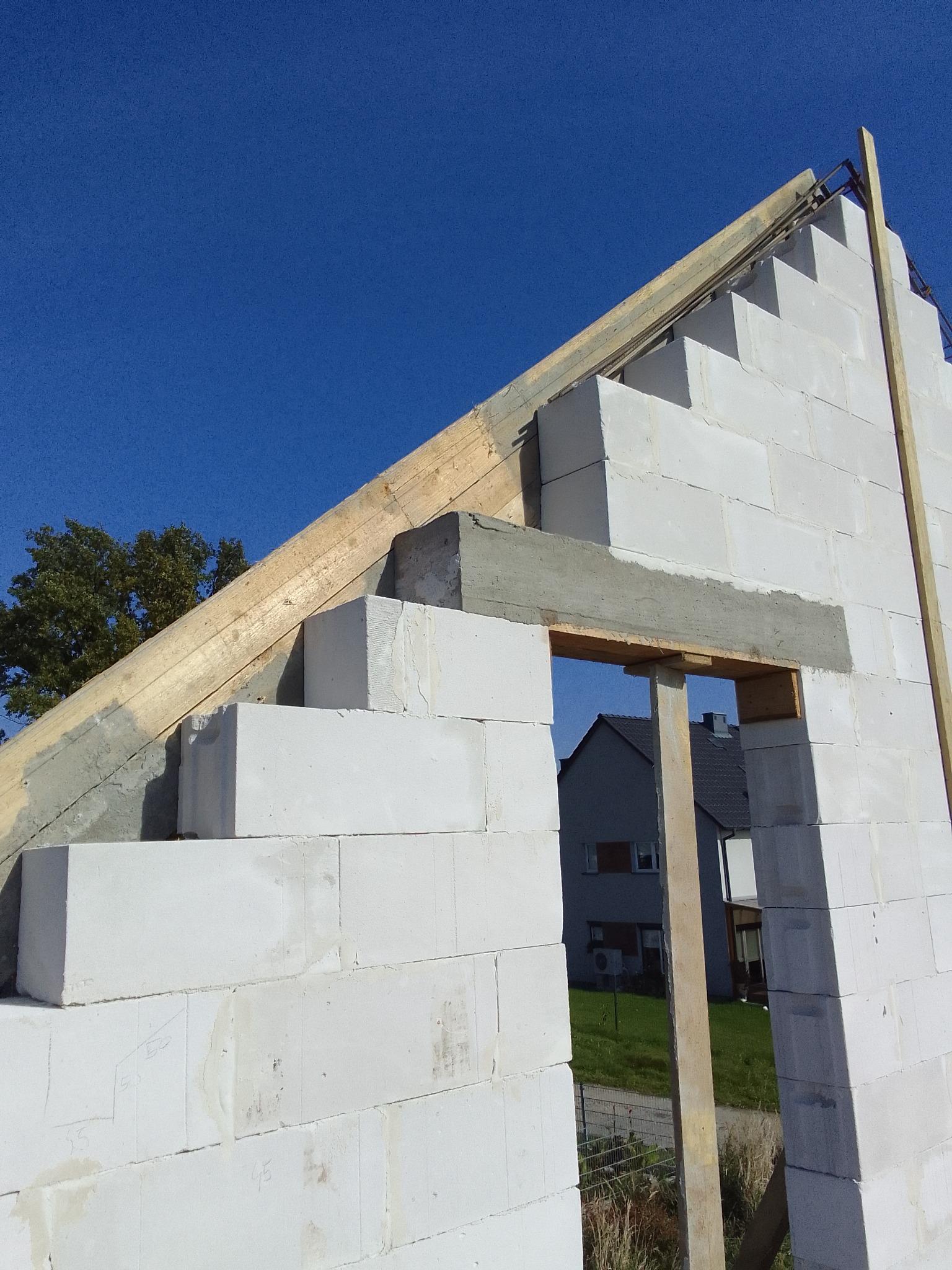 Wznoszenie ścian domu z betonu komórkowego Ytong, widoczne zbrojenie wieńca i otwór okienny z drewnianym szalunkiem, w tle błękitne niebo i fragment sąsiedniego budynku.