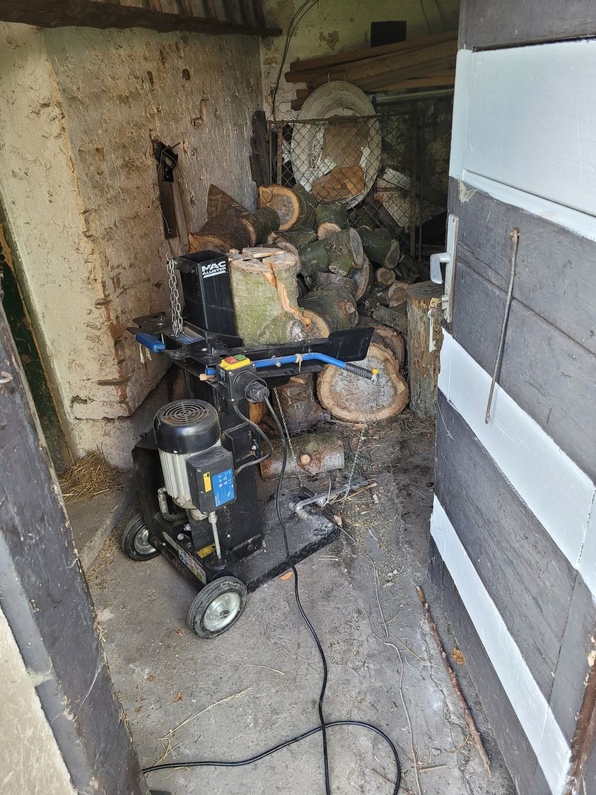 Hydrauliczna łuparka do drewna MAC Allister na kółkach, z łańcuchem zabezpieczającym, stoi w zadaszonym pomieszczeniu z stertą pociętych pni w tle, widoczne ślady użytkowania i zabrudzenia.