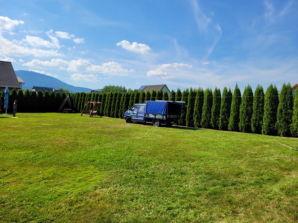 Rozległy trawnik z równo przyciętymi tujami wzdłuż granicy działki, niebieski samochód dostawczy z plandeką zaparkowany obok, huśtawka w tle, widok na góry w oddali.