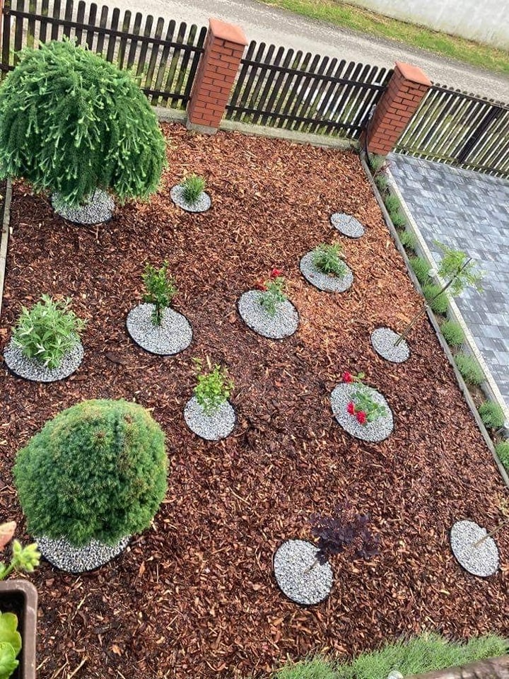 Widok z góry na świeżo założony ogród z młodymi drzewkami i krzewami otoczonymi białym żwirem na tle kory drzewnej, ogrodzenie z ceglanymi słupkami w tle.