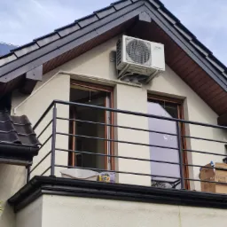 Biała jednostka zewnętrzna klimatyzatora zamontowana na elewacji domu z balkonem, widoczna instalacja odprowadzająca skropliny i okno z naklejką jelenia.