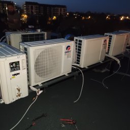 ADEK - Instalacja zewnętrznych jednostek klimatyzacji marki Gree na dachu budynku mieszkalnego, widoczne narzędzia i instalator podczas montażu w warunkach słabego oświetlenia.