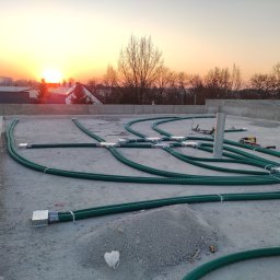 ADEK - Montaż instalacji rekuperacji na dachu budynku, widoczne zielone rury wentylacyjne, złącza, narzędzia i osoba sprawdzająca plany na tle zachodzącego słońca.