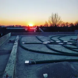 Instalacja wentylacyjna z zielonymi rurami na betonowym dachu podczas zachodu słońca, widok na okoliczne domy i drzewa w tle.