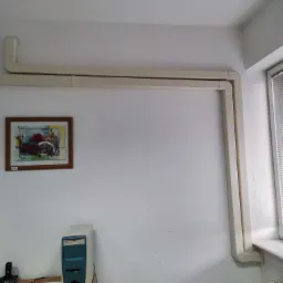 Beżowe kanały wentylacyjne biegnące wzdłuż sufitu i ściany w pomieszczeniu z oknem, obrazem na ścianie i komputerem stacjonarnym.