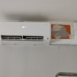 ADEK - Zamontowany na ścianie biały klimatyzator z widocznym wyświetlaczem temperatury, obok niego instalacja maskująca przewody, ślady po naprawach na ścianie.