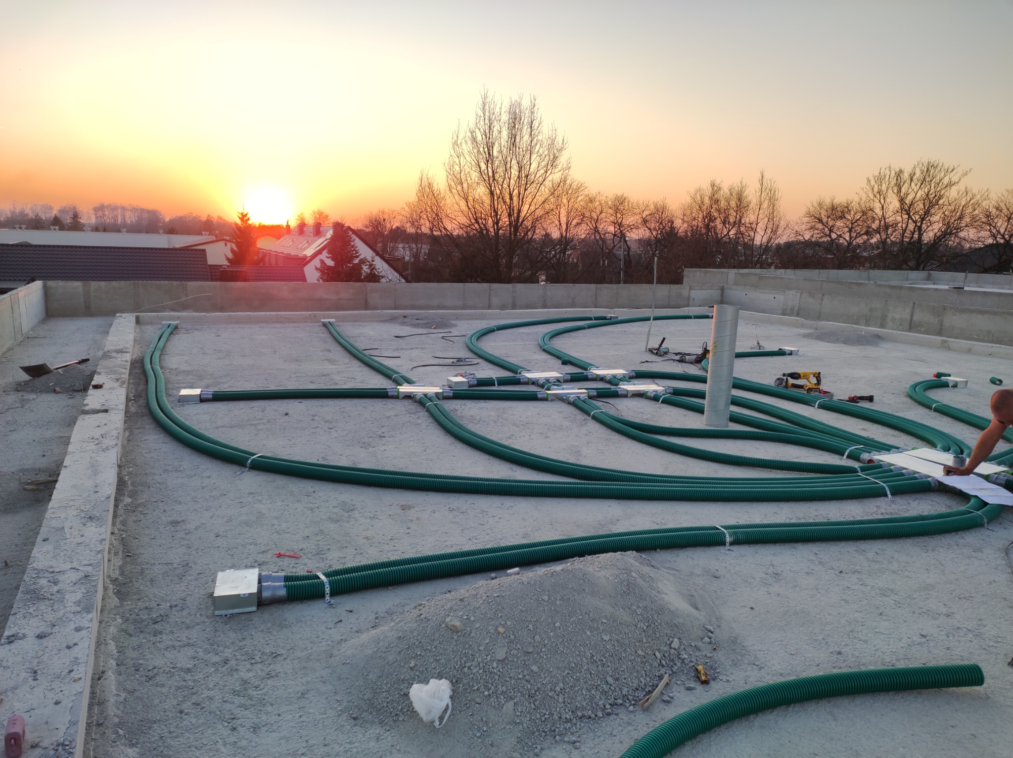 Montaż instalacji rekuperacji na dachu budynku, widoczne zielone rury wentylacyjne, złącza, narzędzia i osoba sprawdzająca plany na tle zachodzącego słońca.