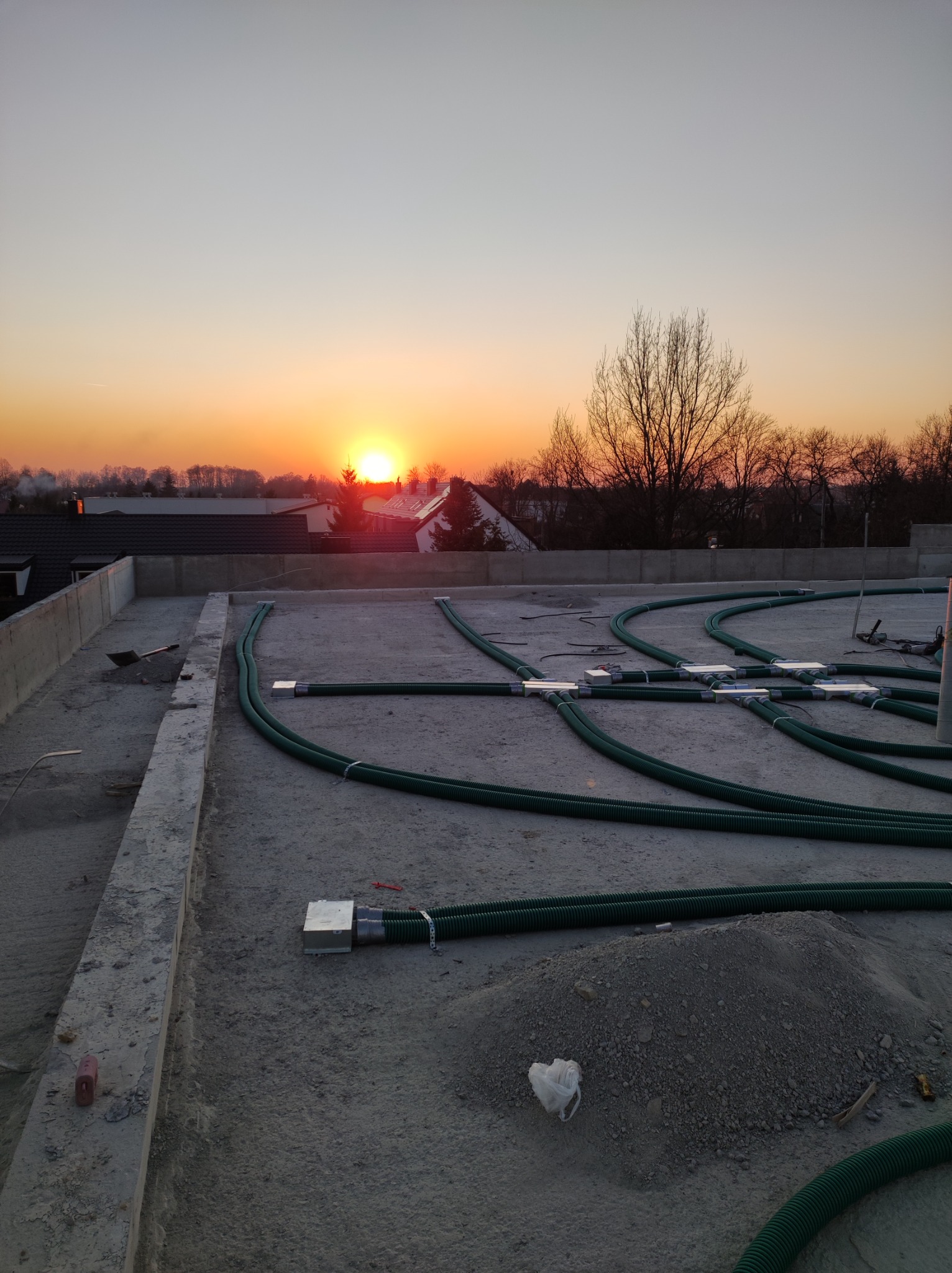 Instalacja rur wentylacyjnych w systemie rekuperacji na betonowym dachu budynku podczas zachodu słońca, widoczne złącza i narzędzia pracy.