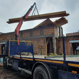 Czerwony żuraw przenosi wiązkę drewnianych belek nad surową konstrukcją domu z cegły, widoczny transport ciężarowy z niebieską platformą, w tle las i pochmurne niebo.