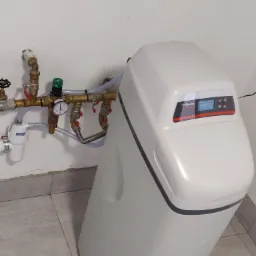 Biały zmiękczacz wody Viessmann podłączony do instalacji hydraulicznej z zaworami, manometrem i filtrem na tle białej ściany i szarych płytek podłogowych.