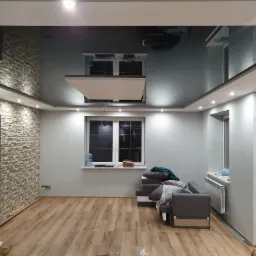 Pokój po remoncie z błyszczącym, ciemnym sufitem napinanym, fragmentem ściany z kamienia dekoracyjnego, drewnianą podłogą i meblami w szarych tonacjach.