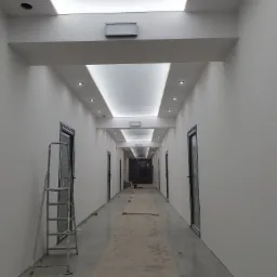 Długi, jasny korytarz w trakcie remontu, widoczne białe ściany, nowe drzwi z niebieską ramą, podwieszany sufit z oświetleniem punktowym i centralnym, drabina oparta o ścianę, podłoga zabezpieczona...