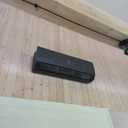 Czarny klimatyzator Haier zamontowany na drewnianej ścianie, z wyświetlaczem temperatury ustawionym na 24 stopnie, widoczny drewniany strop z czarnym oświetleniem szynowym.