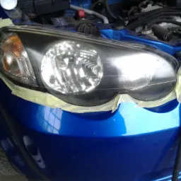 Niebieski samochód z reflektorem zabezpieczonym taśmą maskującą przed polerowaniem lub drobną naprawą lakierniczą, widoczny fragment opony i otwarty silnik.