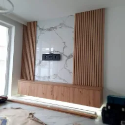 Wykończona ściana telewizyjna z marmurowym panelem, drewnianymi lamelami po bokach i niską szafką RTV z oświetleniem LED u dołu, widoczne narzędzia i zabezpieczenia podłogi.