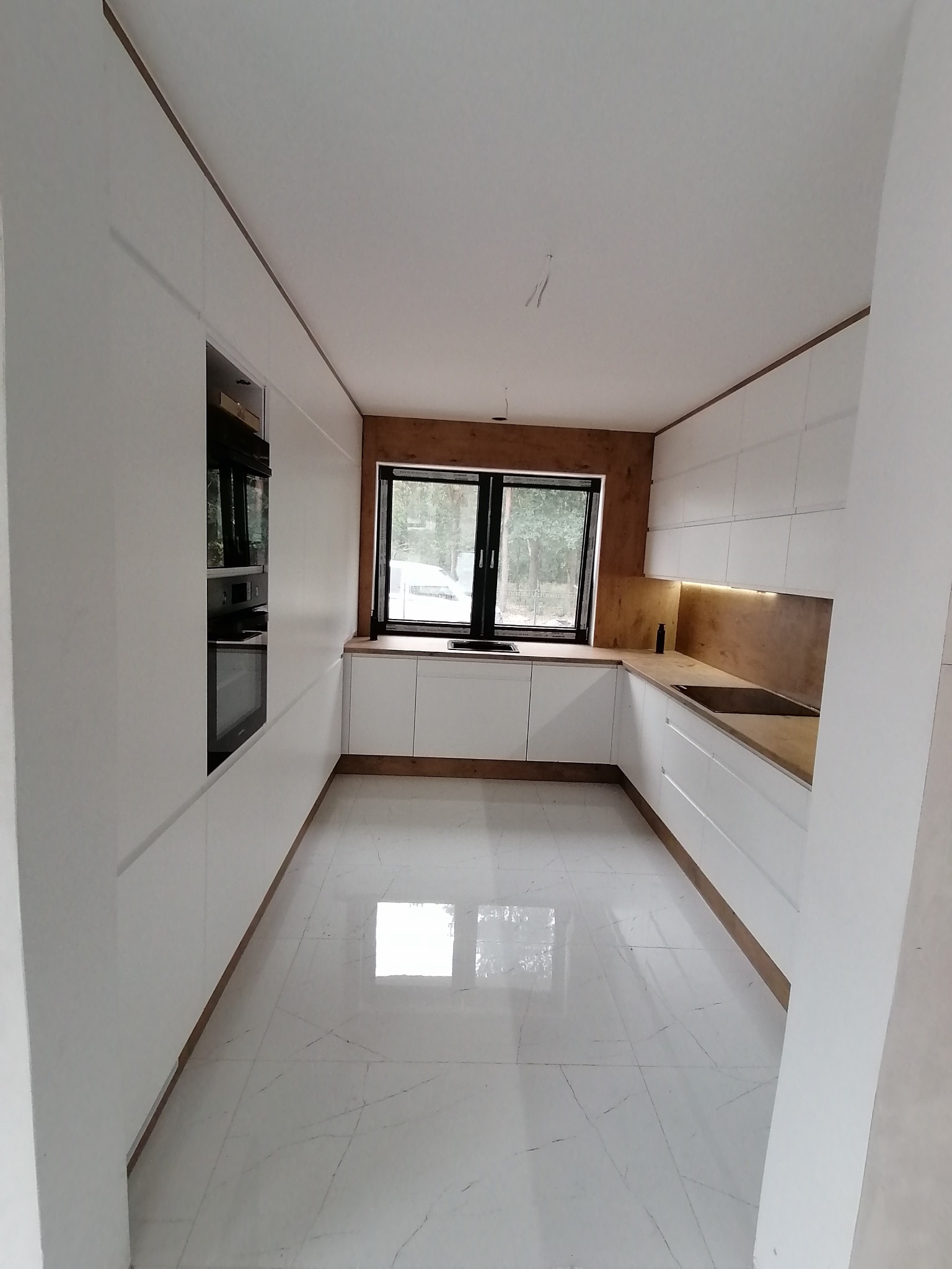 Nowoczesna kuchnia z białymi, minimalistycznymi szafkami, drewnianymi blatami i akcentami w kolorze drewna, widok na okno z widokiem na zieleń, marmurowa podłoga z wyraźnym połyskiem.