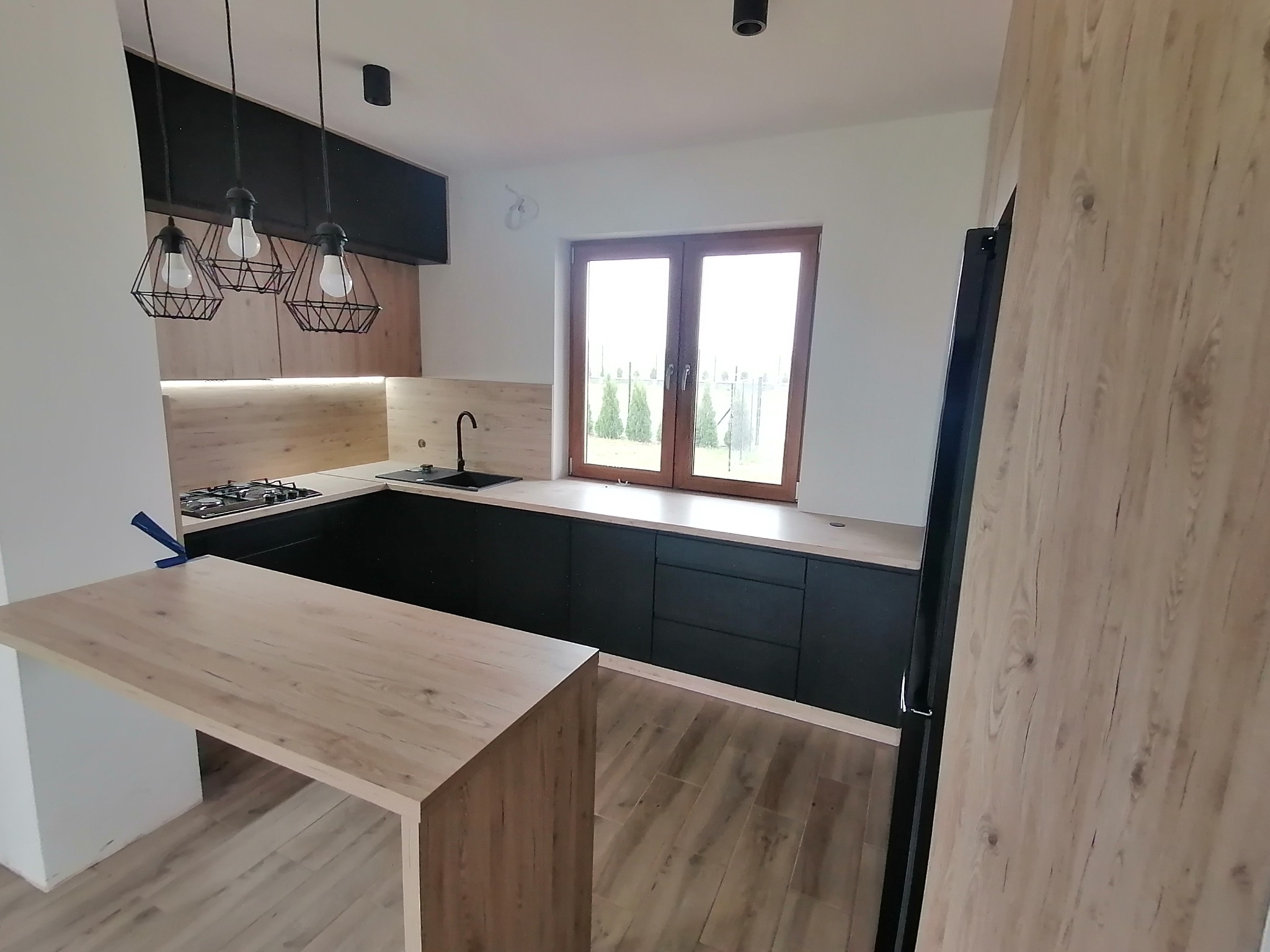 Nowoczesna kuchnia z czarnymi szafkami i drewnianym blatem, oświetlona trzema lampami wiszącymi w stylu industrialnym, z widokiem na ogród przez okno.