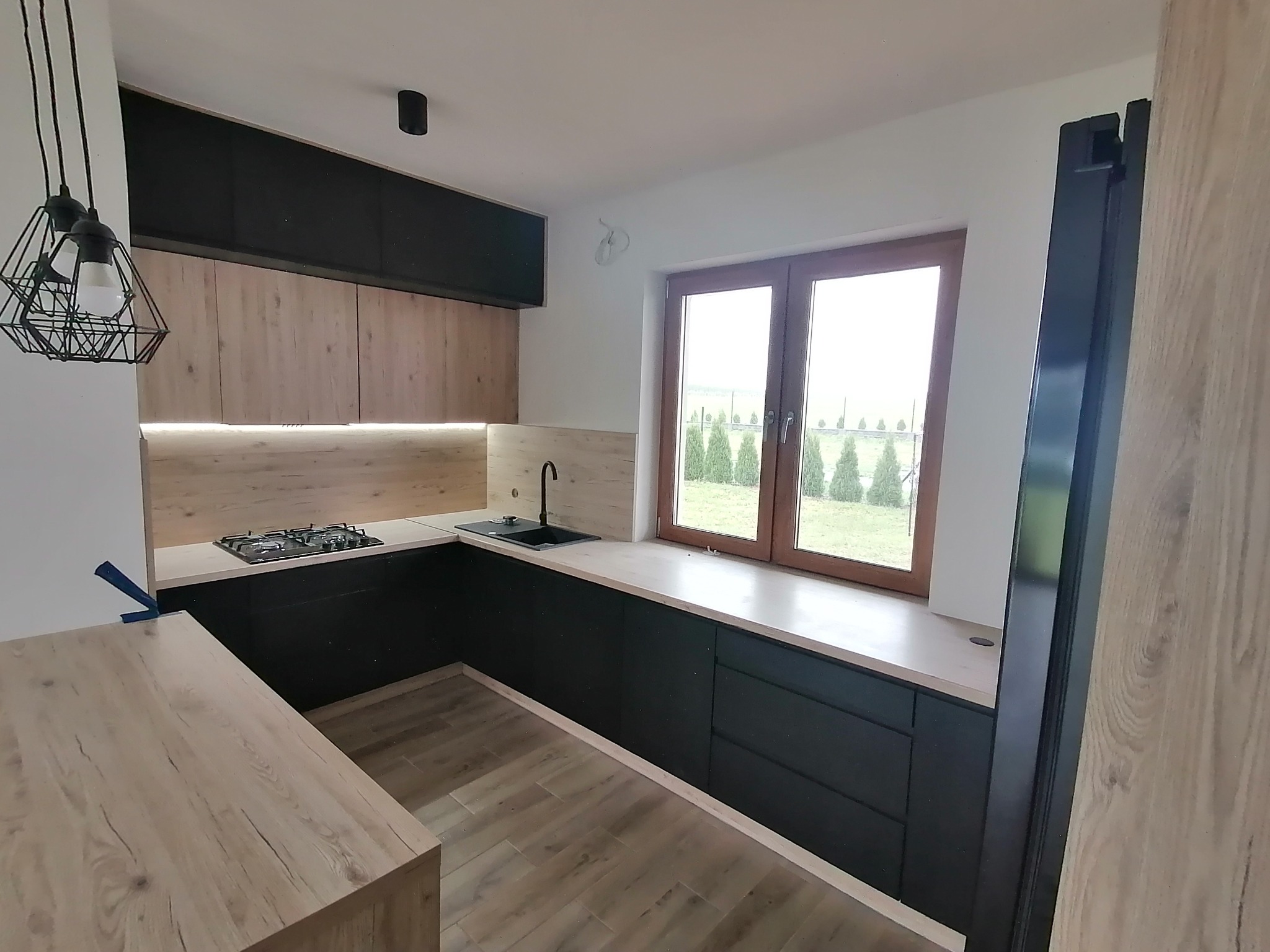 Nowoczesna kuchnia z czarnymi szafkami dolnymi i drewnopodobnymi górnymi, oświetleniem LED podszafkowym, blatem pod oknem i lampami wiszącymi w stylu industrialnym.