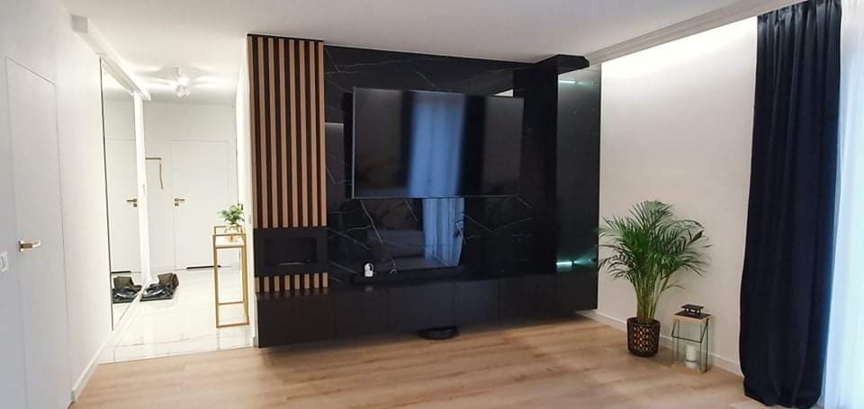 Nowoczesny salon z czarną ścianą telewizyjną z marmurowym wzorem, drewnianymi lamelami i wiszącą szafką RTV, uzupełniony przez rośliny doniczkowe i minimalistyczne dekoracje.