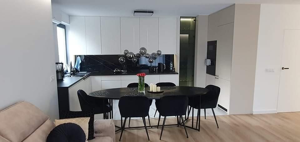 Nowoczesna kuchnia z białymi szafkami, czarnym blatem i panelem, oświetlona dekoracyjną lampą kulistą, z owalnym stołem i czarnymi krzesłami.