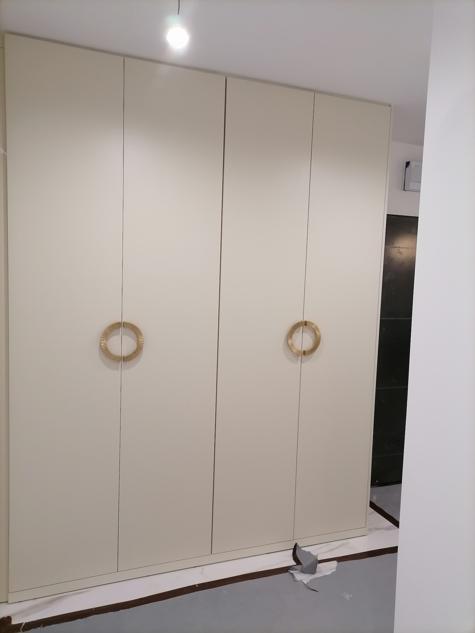 Biała, czterodrzwiowa szafa z okrągłymi, złotymi uchwytami. Widoczne niedociągnięcia wykończeniowe na podłodze.