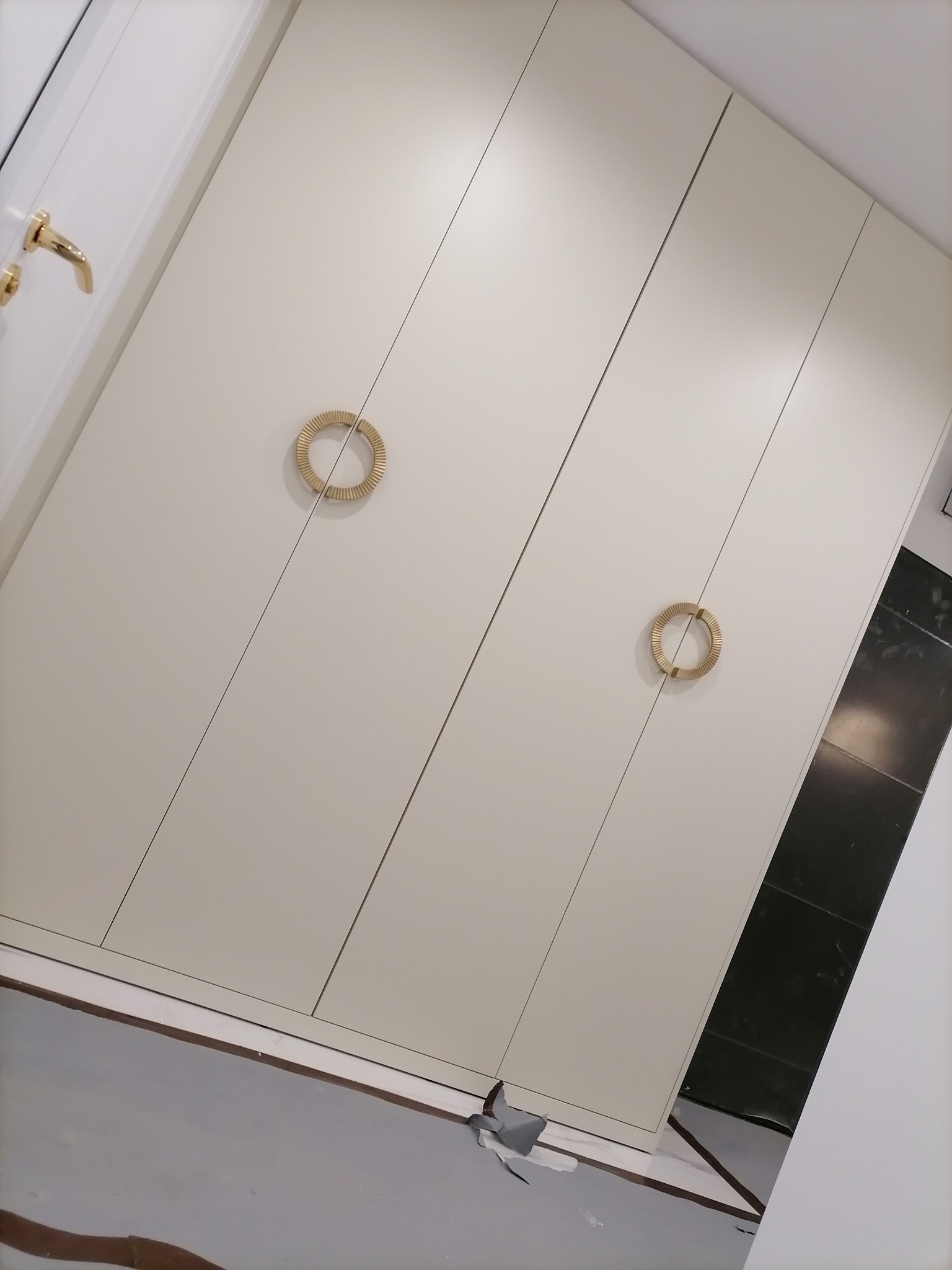 Biała szafa wnękowa z czterema drzwiami, ozdobnymi okrągłymi uchwytami w kolorze złotym, widoczne fragmenty podłogi i ściany.