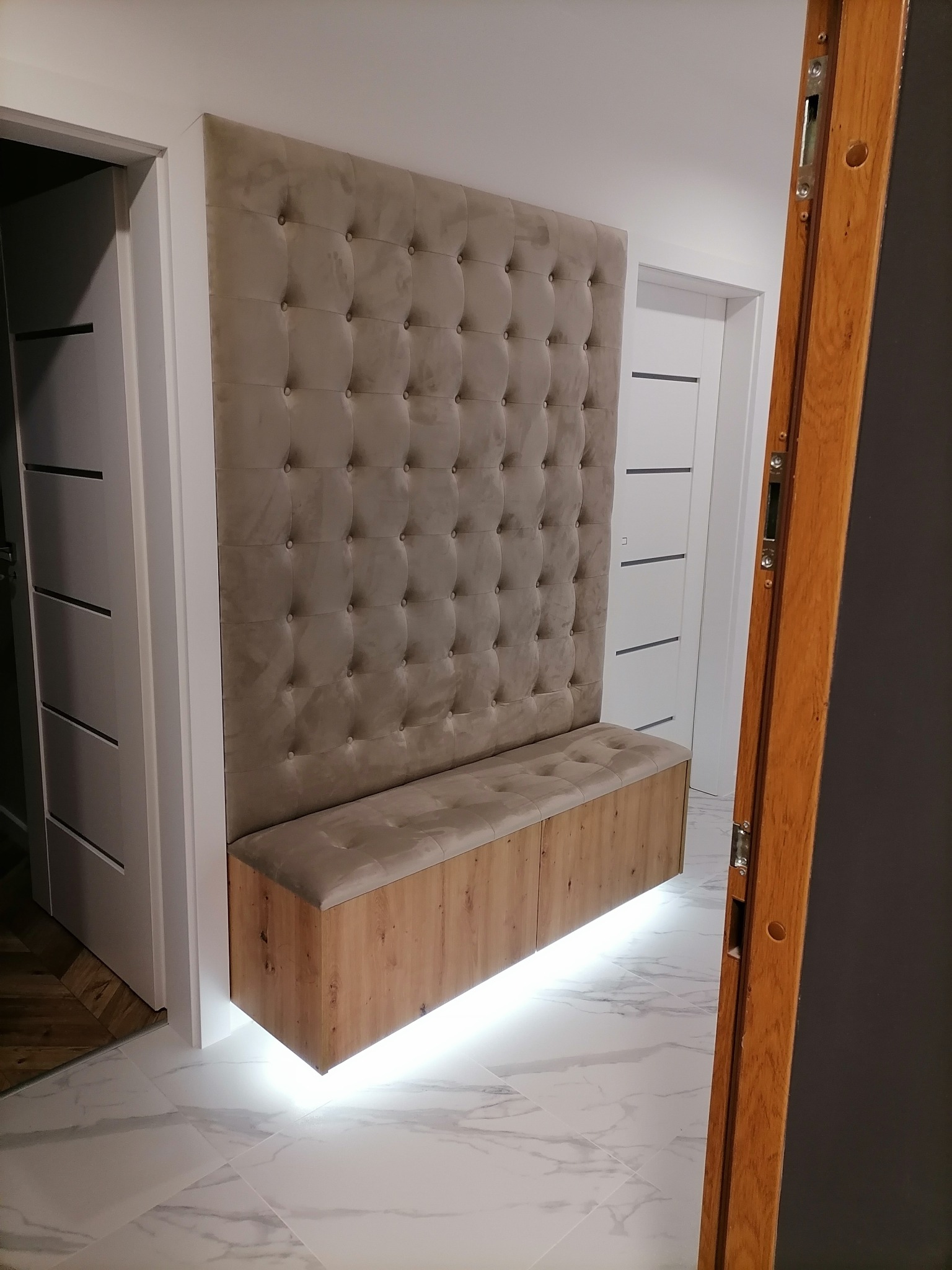 Beżowy, tapicerowany panel ścienny z guzikami, połączony z drewnianą szafką z siedziskiem, podświetlony od dołu, w przedpokoju z marmurową podłogą.