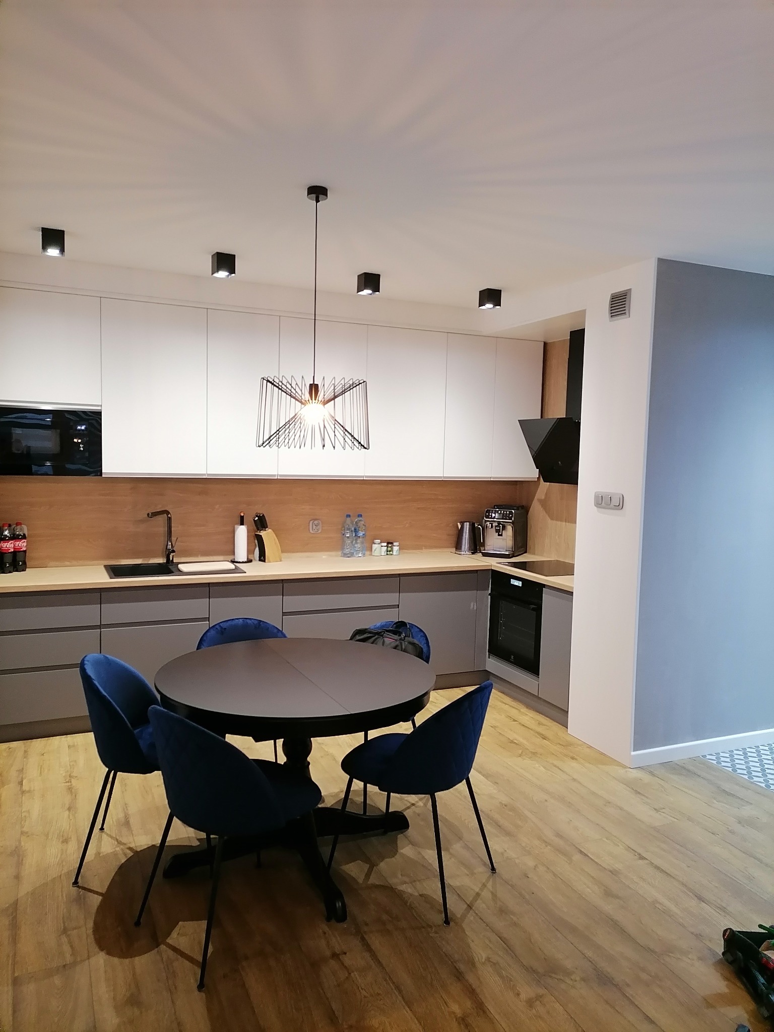 Nowoczesna kuchnia z białymi górnymi szafkami i szarymi dolnymi, drewnianym blatem i okrągłym stołem z granatowymi krzesłami, oświetlona designerską lampą wiszącą.