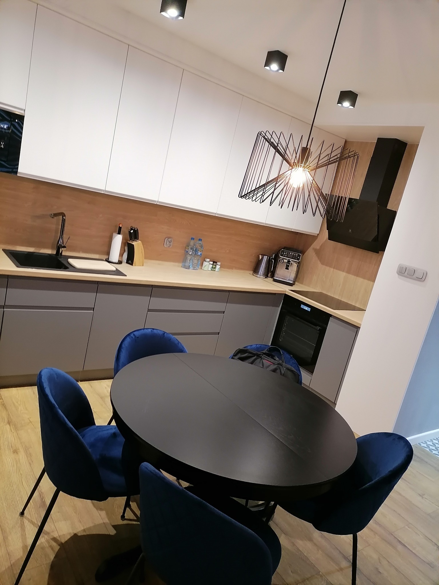 Nowoczesna kuchnia z szarymi szafkami, drewnianym blatem i okrągłym, czarnym stołem otoczonym niebieskimi, welurowymi krzesłami. Nad stołem designerska lampa z metalowymi elementami.