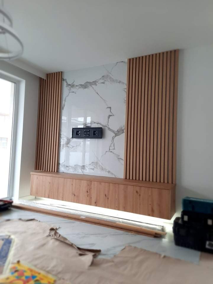 Wykończona ściana telewizyjna z marmurowym panelem, drewnianymi lamelami po bokach i niską szafką RTV z oświetleniem LED u dołu, widoczne narzędzia i zabezpieczenia podłogi.