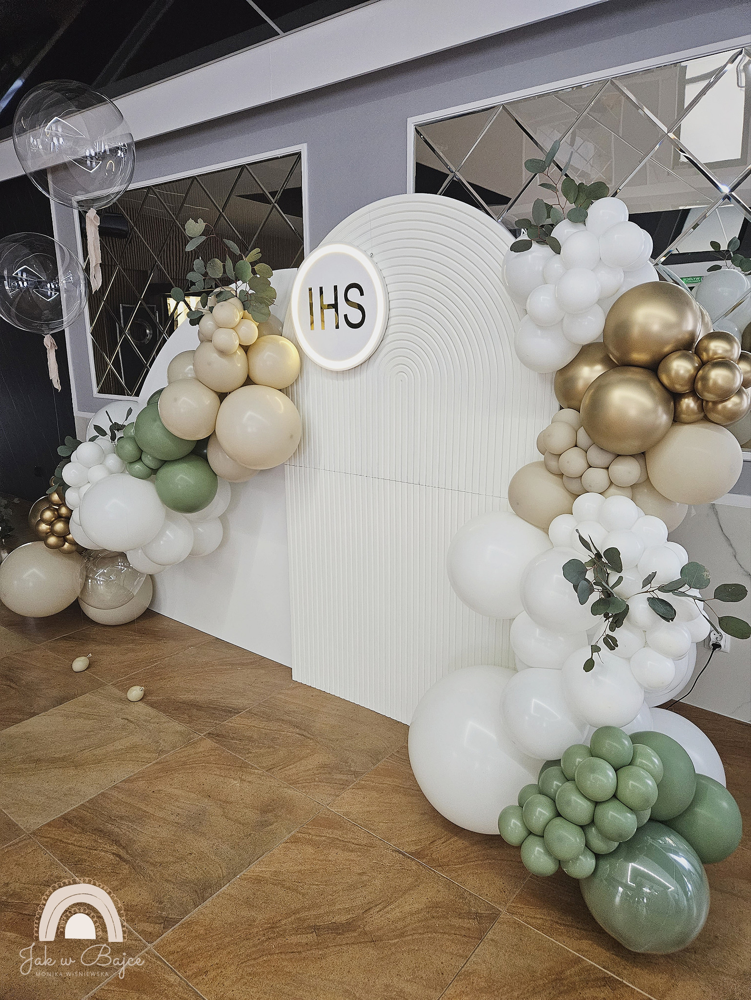 Dekoracja balonowa w odcieniach bieli, beżu, złota i zieleni, z motywem IHS, na tle białej ścianki z geometrycznym wzorem i lustrzanymi elementami, prawdopodobnie na Pierwszą Komunię Świętą.