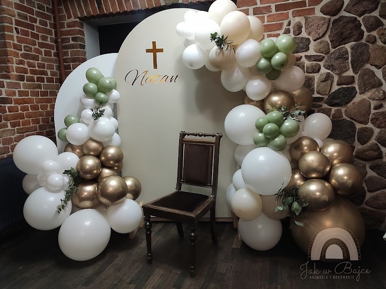 Dekoracja balonowa w odcieniach bieli, złota i zieleni, z drewnianym krzesłem pośrodku i białym panelem z napisem i krzyżem w tle, na tle kamiennej ściany.