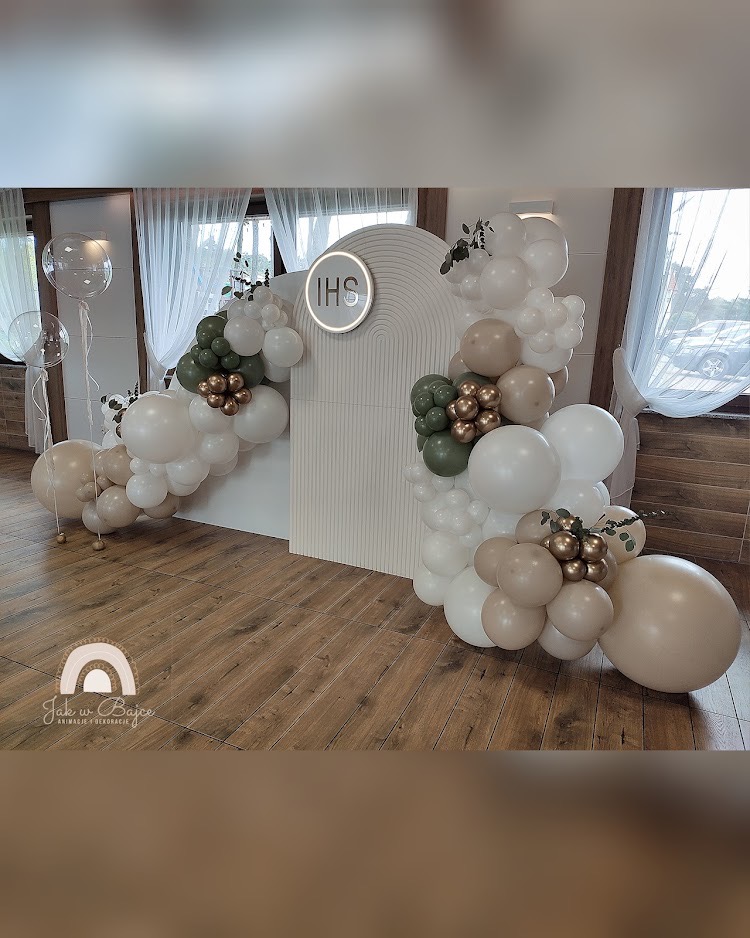 Dekoracja balonowa w odcieniach bieli, beżu i zieleni, z logo IHS w okręgu, na tle białej ścianki z żłobieniami, w jasnym wnętrzu.